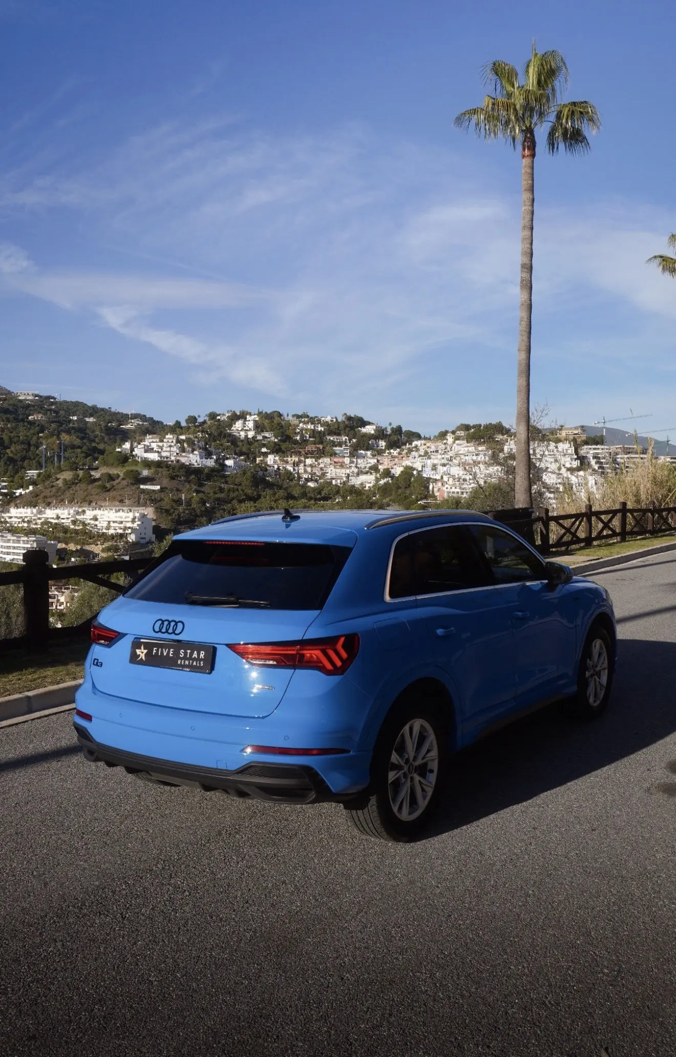 Vista 3/4 trasera del Audi Q3 Turbo Blue con matrícula Five Star Rentals, faros LED dinámicos y pueblo blanco en la colina de Marbella de fondo