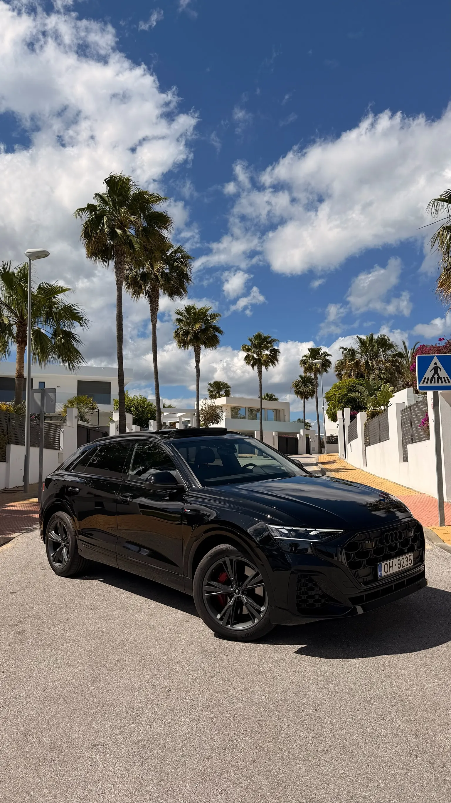 Audi Q8 55 TFSI Quattro S Line in Orca Black metallic, front three-quarter view with panoramic roof open, Marbella palm trees and modern villas