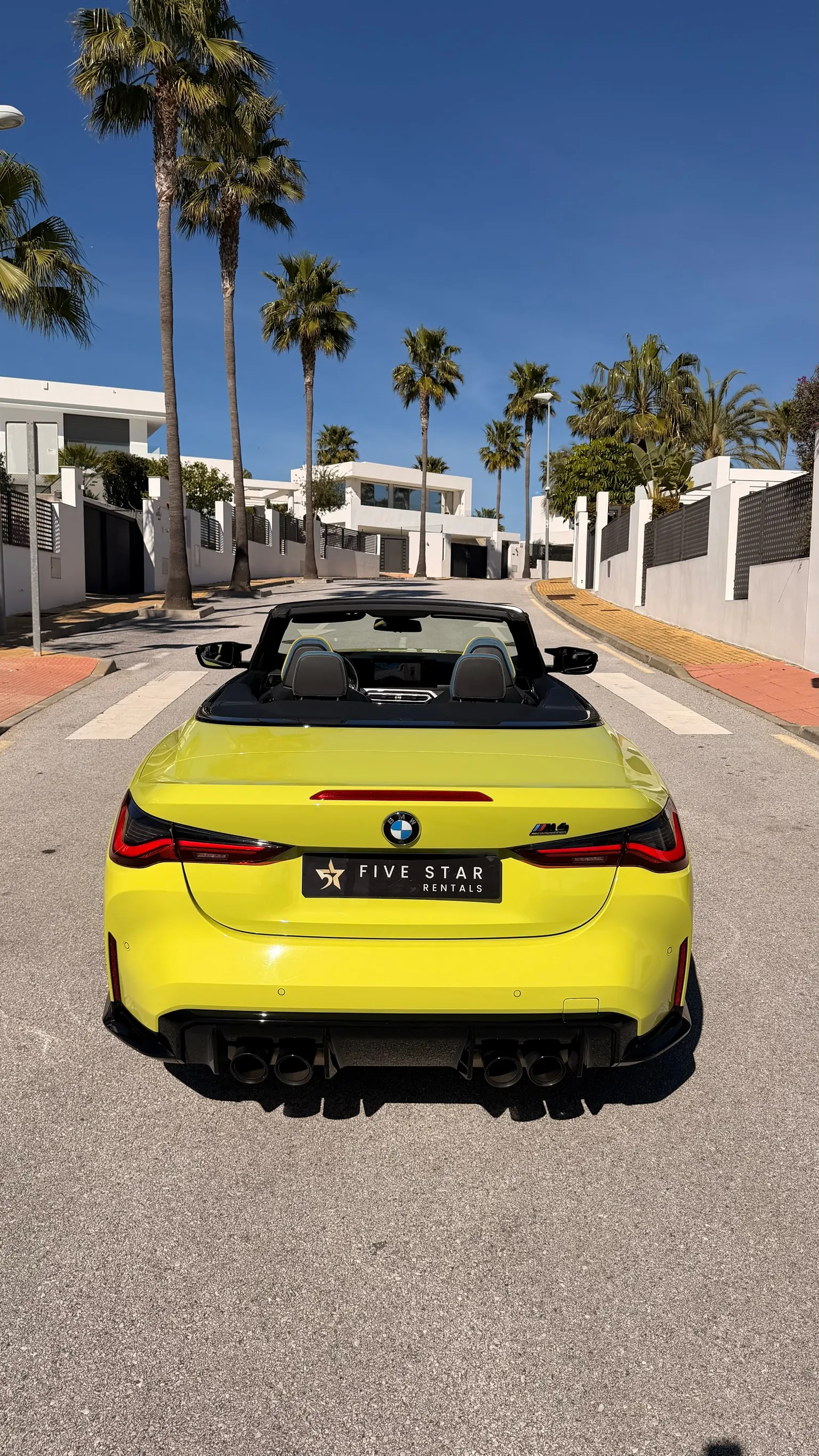 Vista trasera centrada del BMW M4 Competition Cabriolet amarillo con cuádruple escape, insignia M4 Competition y matrícula Five Star Rentals
