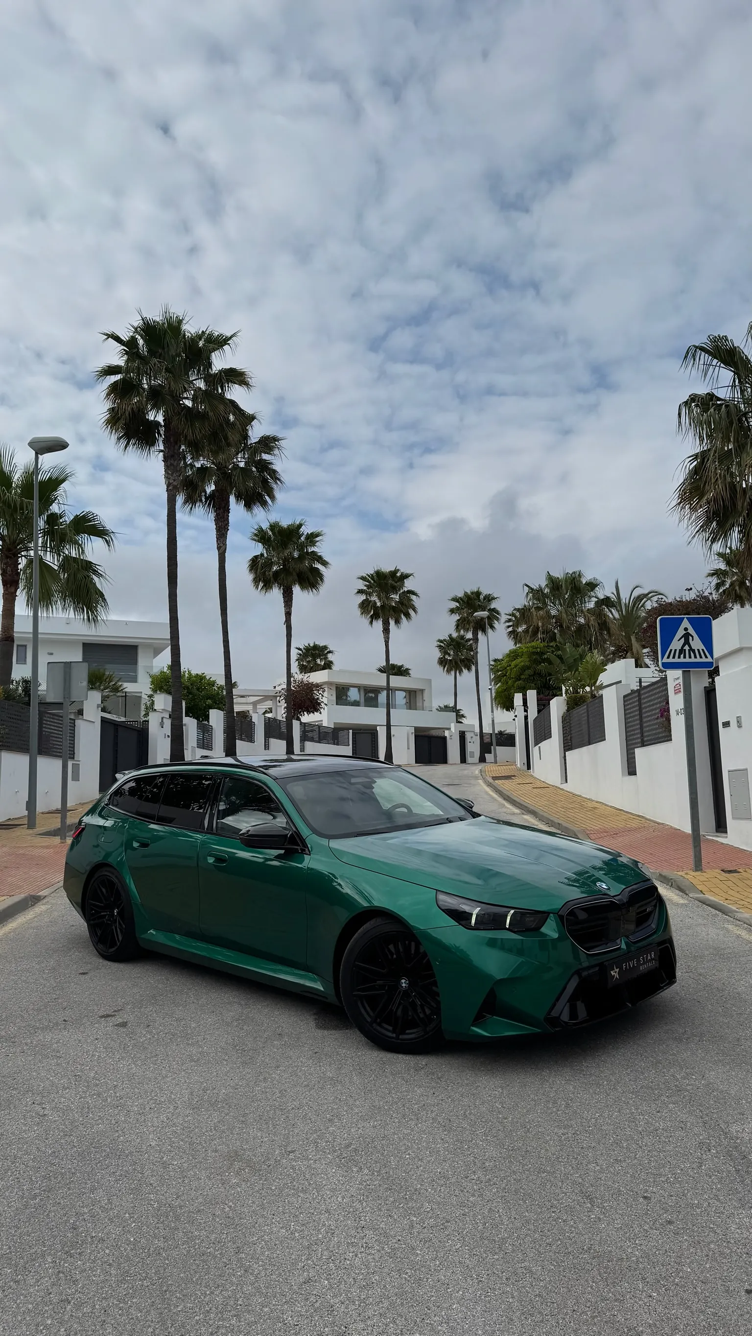 BMW M5 Touring G99 in British Racing Green, front three-quarter view with Marbella palm trees backdrop