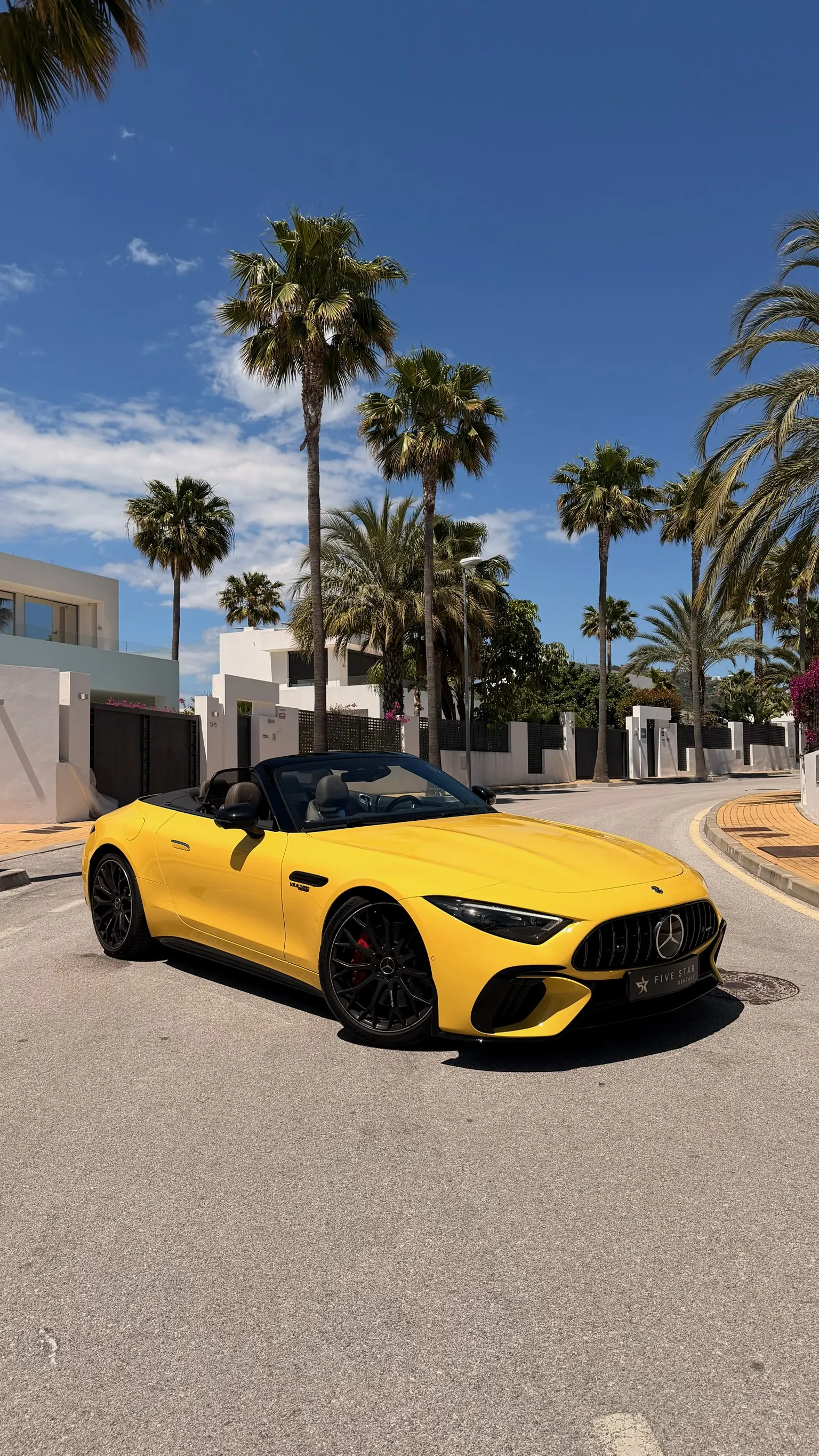 Mercedes-AMG SL 55 4MATIC+ Roadster Sun Yellow con capota abierta, vista 3/4 frontal con matrícula Five Star Rentals, parrilla vertical cromada y palmeras de Marbella