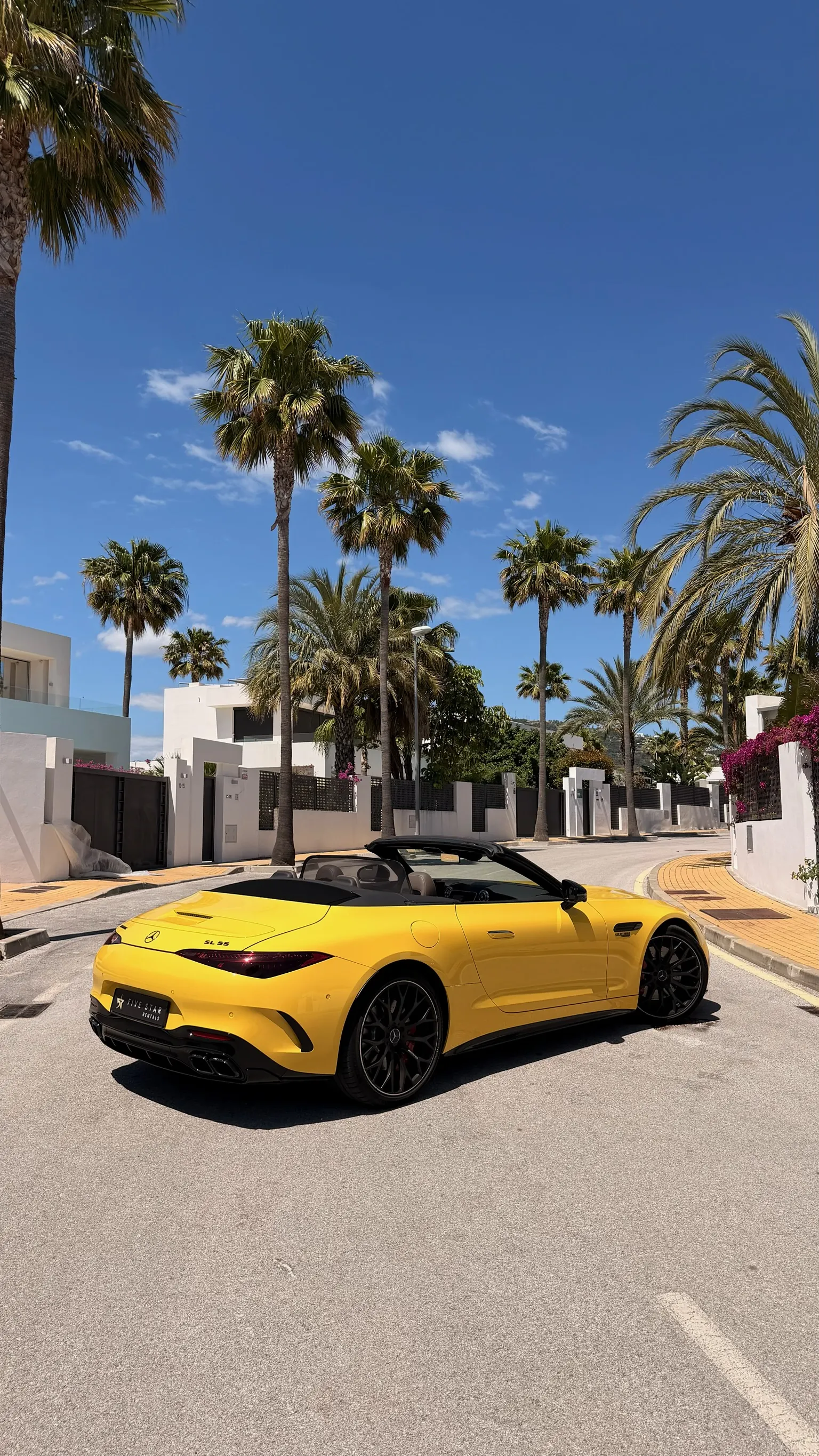 Vista 3/4 trasera del Mercedes-AMG SL 55 Sun Yellow con capota abierta, matrícula Five Star Rentals, palmeras y buganvillas de Marbella de fondo