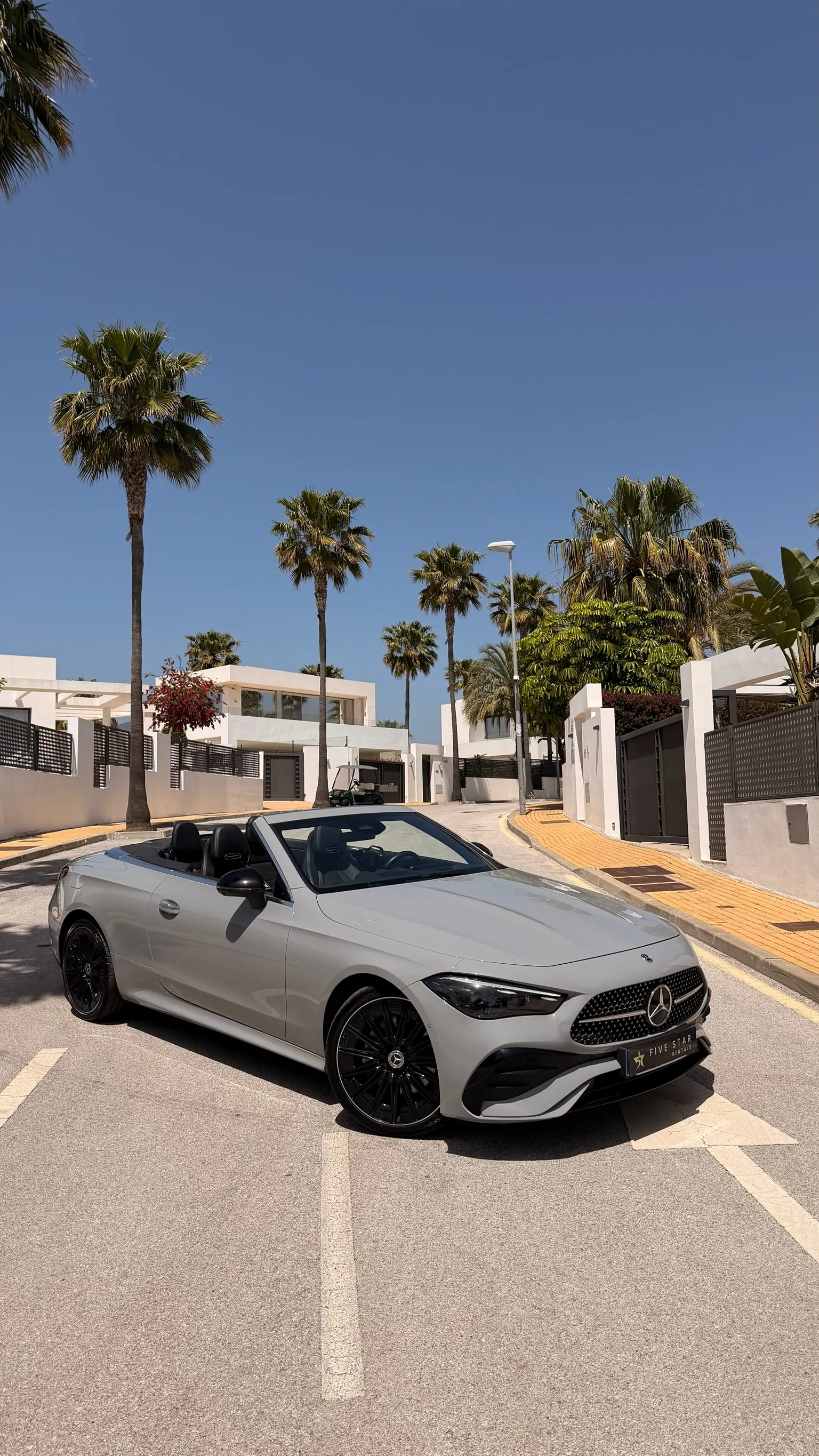 Mercedes-Benz CLE 200 Cabriolet AMG Line gris alpino, vista 3/4 delantera capota abierta con matrícula Five Star Rentals y palmeras de Marbella