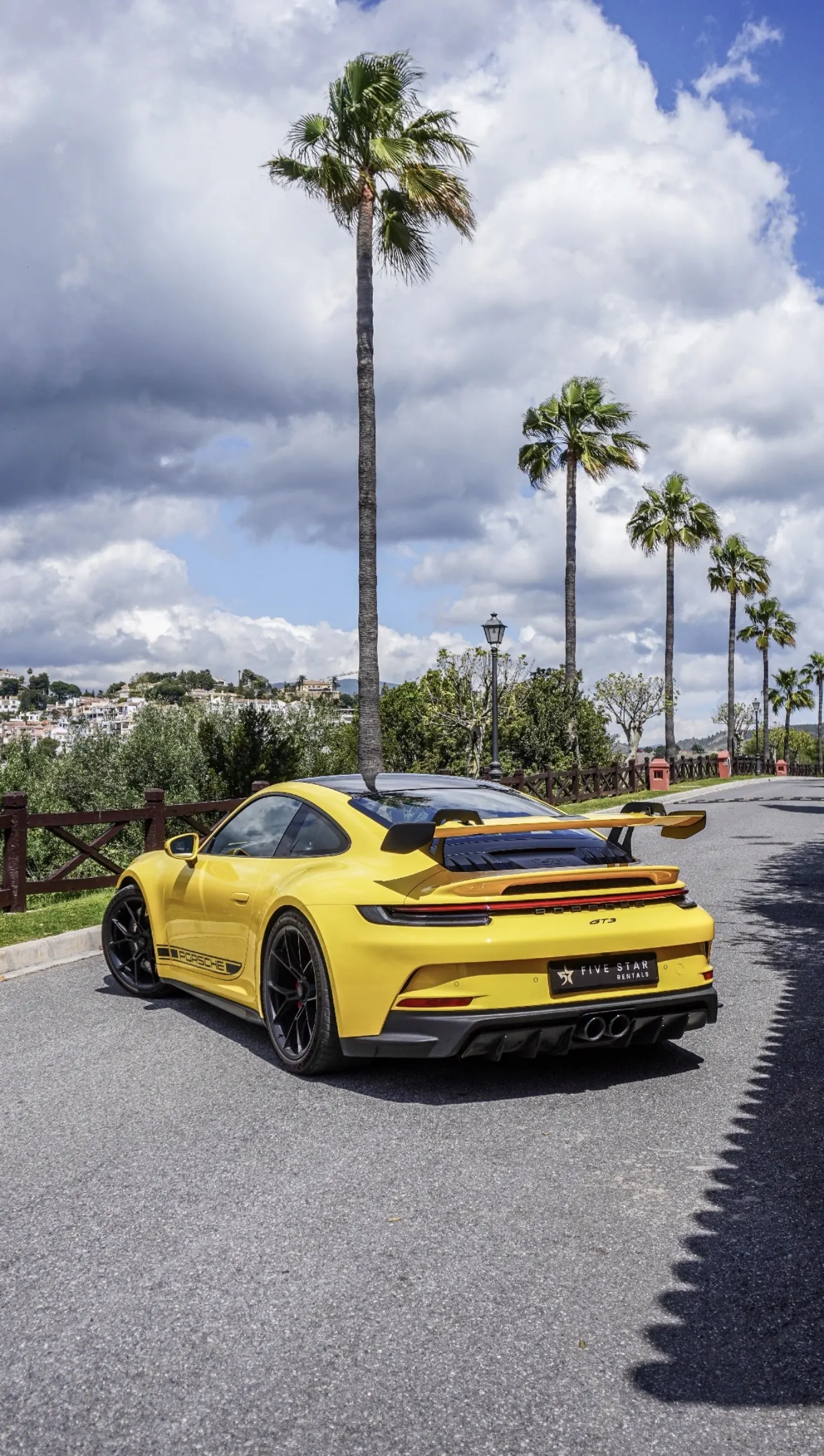 Vista 3/4 trasera del Porsche 911 GT3 992 Racing Yellow con alerón trasero col de cisne, insignia GT3 y matrícula Five Star Rentals bajo las palmeras de Marbella